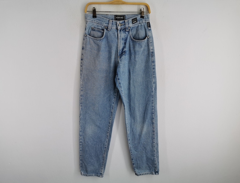May include: Light blue denim jeans with a classic five-pocket design. The jeans are hanging on a wooden hanger against a white background. The brand name "VERSACE" is visible on the waistband.