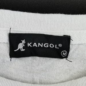 Pu&ograve; includere: Primo piano di una felpa bianca con etichetta nera. L'etichetta mostra il marchio "KANGOL" in bianco, un logo canguro e l'indicazione della taglia "M".
