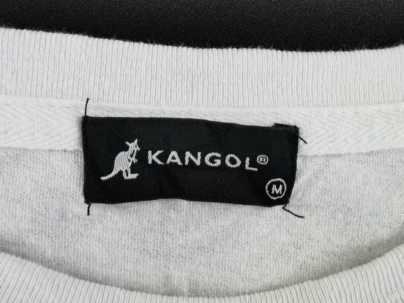 Pu&ograve; includere: Primo piano di una felpa bianca con etichetta nera. L'etichetta mostra il marchio "KANGOL" in bianco, un logo canguro e l'indicazione della taglia "M".