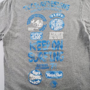 May include: Gray t-shirt with blue graphics. The shirt features text that says "Keep on Surfing" and other phrases like "Captures the Spirit of Surfing" and "Everybody Loves Sunshine". The shirt has short sleeves.