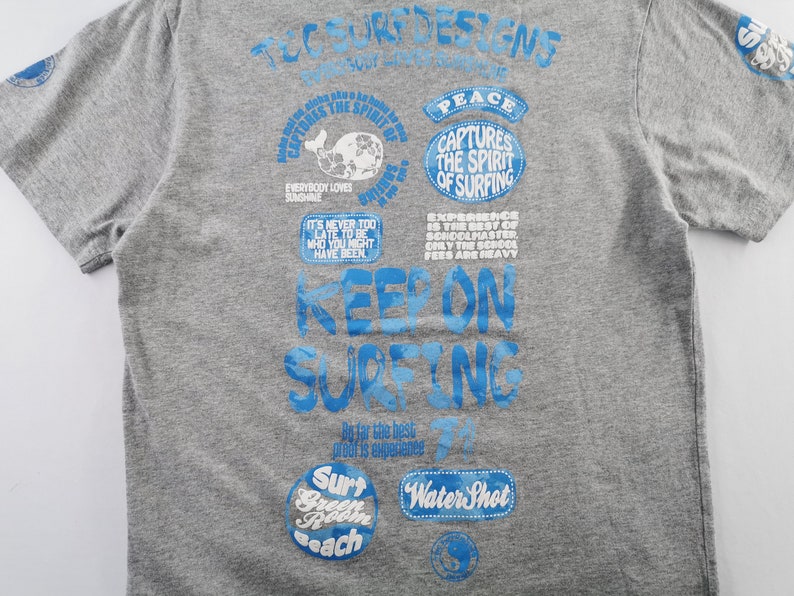 May include: Gray t-shirt with blue graphics. The shirt features text that says "Keep on Surfing" and other phrases like "Captures the Spirit of Surfing" and "Everybody Loves Sunshine". The shirt has short sleeves.