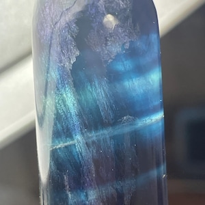 May include: A polished, cylindrical crystal tower featuring deep blue and violet hues. The translucent stone displays internal banding, fibrous inclusions, and natural fractures that create a layered, ethereal appearance throughout the structure.