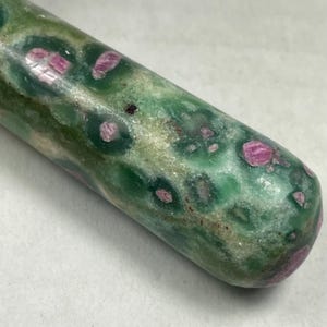 May include: A cylindrical polished stone wand featuring a mottled green base with embedded deep pink and magenta crystalline inclusions. The surface has a smooth, glossy texture with natural variations in color and mineral patterns throughout.