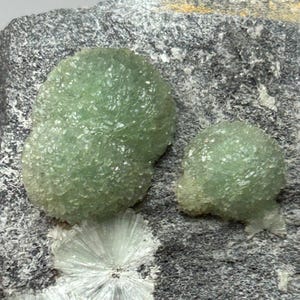 May include: Two rounded, light green, crystalline mineral formations sit atop a dark gray, textured rock matrix. Below the green clusters, a small, white, fan-shaped mineral spray is visible, contrasting with the dark, rough stone surface.