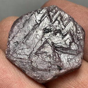 May include: A raw, hexagonal-shaped ruby crystal with a muted purplish-red hue. The surface features a complex network of intersecting geometric striations, natural etchings, and small dark inclusions across its flat, crystalline face.