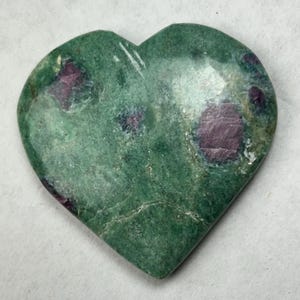 May include: A heart-shaped polished stone featuring a mottled green base color with several embedded deep magenta and dark purple inclusions throughout the surface. The stone has a smooth, glossy texture and a distinct heart silhouette with a pointed base.