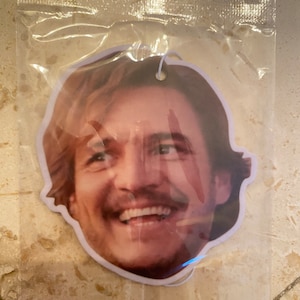May include: A white car air freshener with a printed image of a man's face smiling.