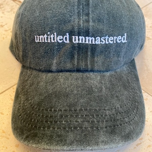 May include: A dark gray baseball cap with the words "untitled unmastered" embroidered in white. The cap has a curved brim and a slightly faded, textured appearance, suggesting a vintage or worn look.