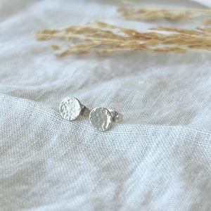May include: A pair of small, hammered silver stud earrings. The earrings have a textured, circular design. The background is a soft, neutral fabric with a few sprigs of dried grass.