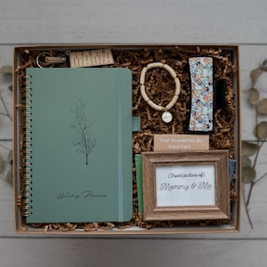 May include: A green gift box filled with items for a new mom. The box includes a journal, a wooden bead bracelet, a small picture frame, and a keychain. The journal has the words "Whisky River" printed on the cover.