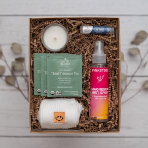 May include: A gift box filled with self-care items for expecting mothers. The box includes a candle, tea bags, a magnesium mist spray, lip balm, and a pair of white socks with a smiley face.