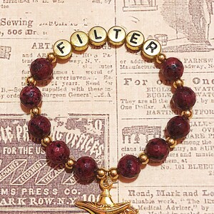 May include: A beaded bracelet with the word "FILTER" spelled out in gold letter beads. The bracelet features dark red speckled beads and a gold-colored genie lamp charm. The background is a vintage newspaper.
