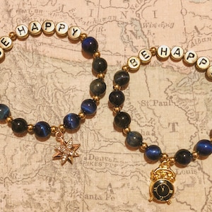 May include: Two beaded bracelets with the words "BE HAPPY" spelled out in white letter beads. The bracelets feature dark blue and black beads, gold accents, and charms, one a starburst and the other a small clock.