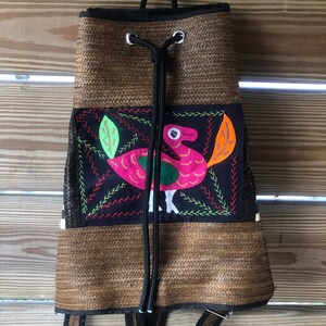 Puede incluir: Una mochila tejida marrón con cierre de cordón negro y correas negras. La parte delantera de la mochila presenta un panel de tela negro con un diseño de pájaro bordado de colores.