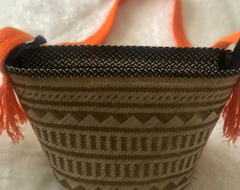 May include: A small woven bag with a geometric pattern in brown and beige. The bag has a bright orange strap with tassels.