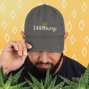 May include: A gray distressed baseball cap with the word "FARMACY" embroidered in white with a small floral design in orange and yellow.
