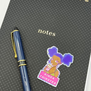 May include: A black notebook with white polka dots and gold spiral binding. A blue pen with gold accents rests on the notebook. A sticker with a cartoon girl drinking boba tea and the text "KINDA THIRSTEA" is also on the notebook.