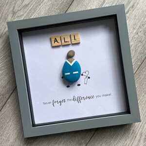 Puede incluir: Una obra de arte enmarcada que presenta a una persona de guijarros con un uniforme azul que sostiene un coraz&oacute;n y un estetoscopio. El nombre "ALI" est&aacute; escrito con fichas de letras encima de la figura. El texto "&iexcl;Nunca olvides la diferencia que haces!" est&aacute; escrito debajo de la figura.