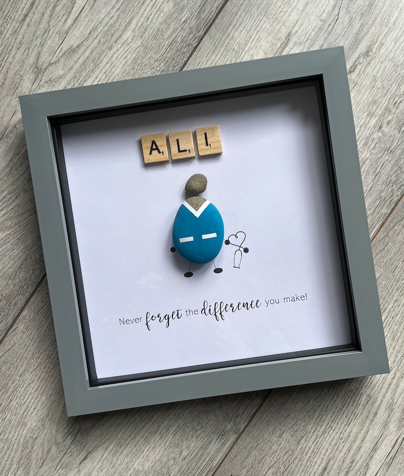 Puede incluir: Una obra de arte enmarcada que presenta a una persona de guijarros con un uniforme azul que sostiene un coraz&oacute;n y un estetoscopio. El nombre "ALI" est&aacute; escrito con fichas de letras encima de la figura. El texto "&iexcl;Nunca olvides la diferencia que haces!" est&aacute; escrito debajo de la figura.