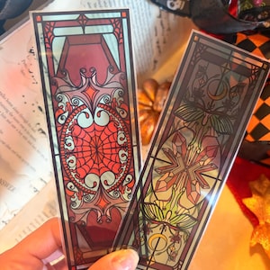 May include: Two rectangular bookmarks featuring stained glass style designs. The left bookmark displays a red spiderweb and coffin motif. The right bookmark features a moth, crystals, and crescent moon imagery in green and red tones.