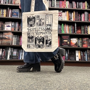 May include: A cream-colored tote bag with the words "HOPELESS ROMANTIC" and various illustrations in black. The bag is held in front of a bookshelf filled with books. The person is wearing black platform boots and blue jeans.