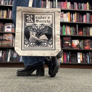 Op de afbeelding: Een witte canvas tote bag met zwarte handvatten en een zwart-wit grafisch ontwerp. De grafiek toont een boek met een hartvorm en de tekst "Reader's Society".