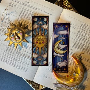 May include: Two celestial-themed bookmarks and two gold-colored ornaments rest on an open book. The bookmarks feature sun and moon designs with stained-glass effects. One ornament is a sun with a smiling face, and the other is a crescent moon with a star.