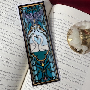 May include: A rectangular bookmark with a stained-glass design. It features two white swans forming a heart shape, surrounded by teal and blue floral patterns. Purple wisteria hangs from the top. The bookmark is on an open book.