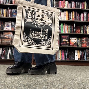May include: A beige tote bag with the words "Book Collector" in a decorative font, featuring a black and white illustration of a bookshelf. The bag is held in front of a bookshelf filled with books. The person is wearing black platform shoes.