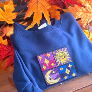 May include: A navy blue crewneck sweatshirt featuring a square embroidered patch on the chest. The patch is divided into four quadrants displaying a yellow sun, a yellow crescent moon, and geometric patterns in shades of purple, pink, and blue.