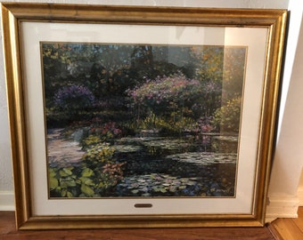 Puede incluir: Pintura de paisaje enmarcada que representa un estanque con nenúfares, rodeado de exuberante vegetación y flores coloridas. La obra de arte está enmarcada en un marco de madera dorado, añadiendo un toque de elegancia a la escena.
