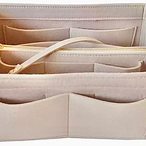 May include: Beige felt handbag organizer with multiple compartments and a zippered pouch. Features a light-colored strap and gold-colored zipper. The organizer has several pockets for storage and organization.