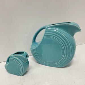 May include: A large turquoise ceramic pitcher and a smaller matching turquoise ceramic creamer, both featuring a rounded shape with concentric circular ring patterns on the sides, a curved handle, and a smooth, glossy glazed finish.