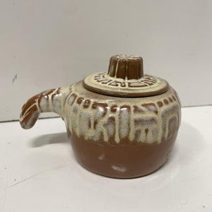 May include: A brown ceramic teapot with a lid and handle. The teapot has a light brown glaze that drips down the sides. The lid has a decorative knob and a lighter brown rim.
