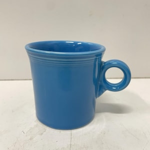 May include: A solid blue ceramic mug featuring a smooth, cylindrical body with a distinct horizontal ring detail near the top rim and a small, circular handle attached to the side. The mug has a glossy finish and a consistent, vibrant blue colour.