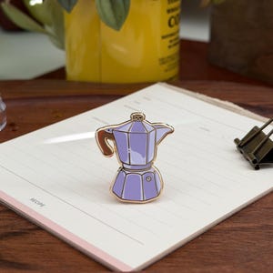 Puede incluir: Un pin de esmalte de color lavanda con forma de cafetera moka. El pin presenta bordes de metal dorado, un asa marrón y un pequeño pomo en la tapa. El diseño incluye un cuerpo de color violeta claro con un detalle de sombra en violeta oscuro.