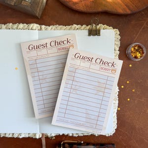 May include: Two rectangular paper guest check pads with a cream background, brown grid lines, and star patterns. The text reads Guest Check, Teddy's Cafe Coffee & Tea, and Thank you - have a great day. Each pad features the number 80897 and a bear icon.