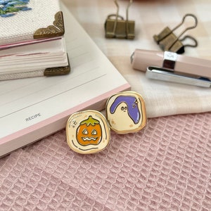 May include: Two gold-colored enamel pins depicting a ghost and a jack-o'-lantern on a white background. The ghost is purple and has a surprised expression. The jack-o'-lantern is orange and has a carved face.