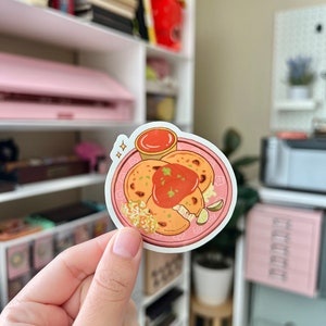 May include: A sticker of a plate of pupusas with a side of salsa. The pupusas are golden brown and have a white filling. The salsa is red and is in a small bowl. The plate is pink and has a white rim.