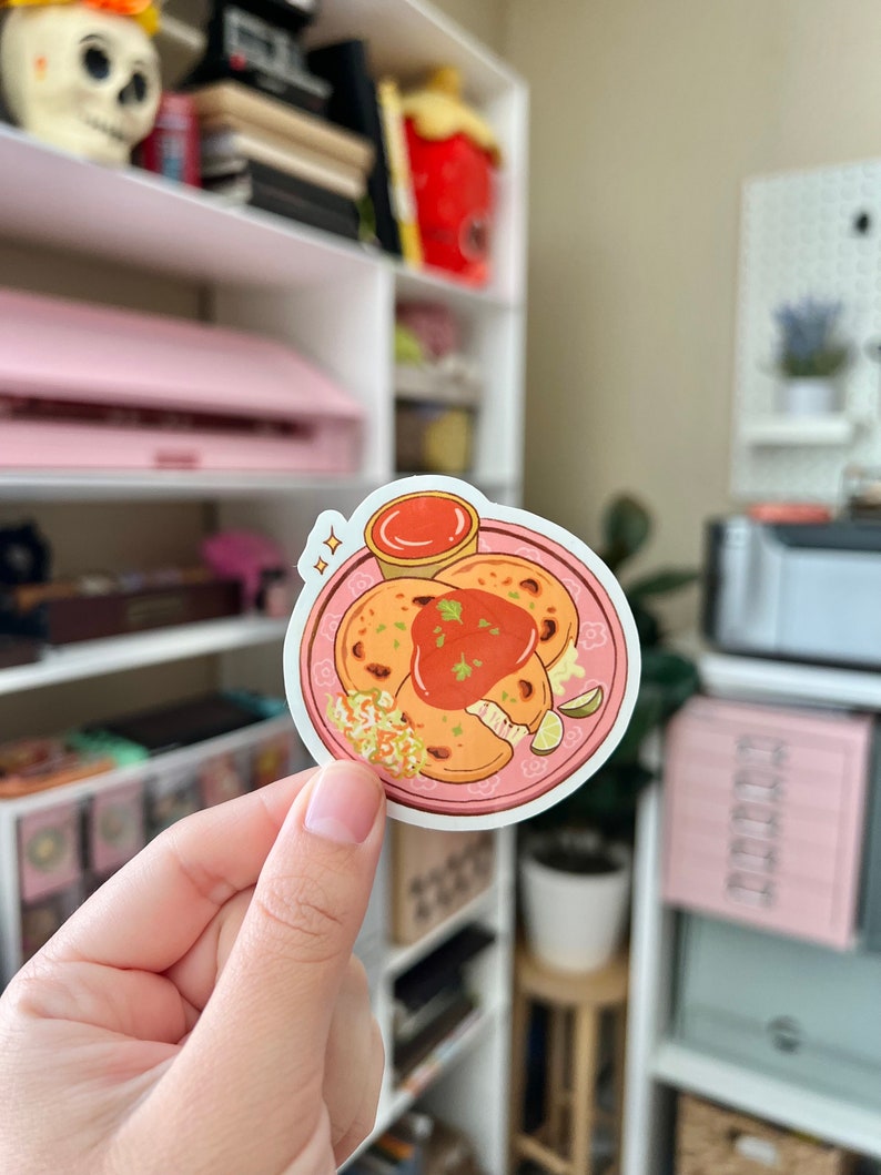 May include: A sticker of a plate of pupusas with a side of salsa. The pupusas are golden brown and have a white filling. The salsa is red and is in a small bowl. The plate is pink and has a white rim.