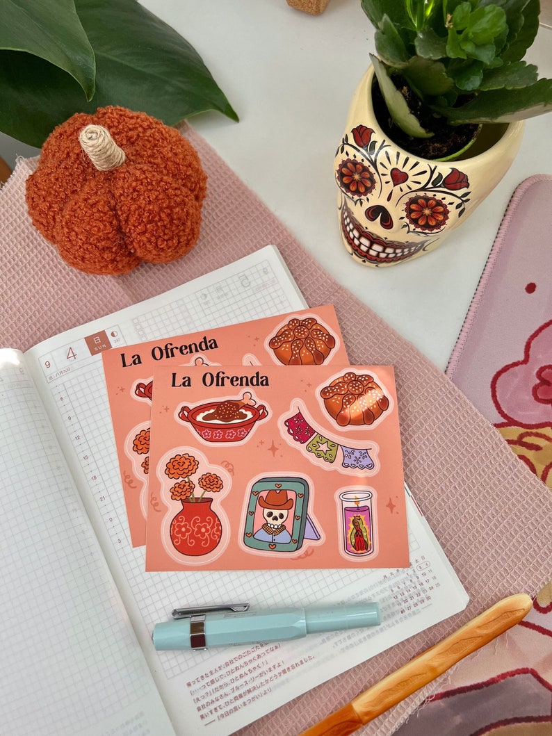 Op de afbeelding: Twee vellen stickers met een thema van D&iacute;a de Muertos. De stickers bevatten een schedel, bloemen, eten en kaarsen. De tekst op de stickers luidt "La Ofrenda".