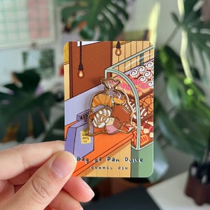 K&ouml;nnte beinhalten: Eine goldene Emaille-Anstecknadel, die einen braunen Papierbeutel zeigt, der mit Pan Dulce gef&uuml;llt ist, einer Art s&uuml;&szlig;em Brot. Die Anstecknadel ist vor einem Comic-Hintergrund einer B&auml;ckerei mit einer Kasse und Regalen mit Geb&auml;ck platziert. Der Text "Bag of Pan Dulce Enamel Pin" ist unter der Anstecknadel gedruckt.