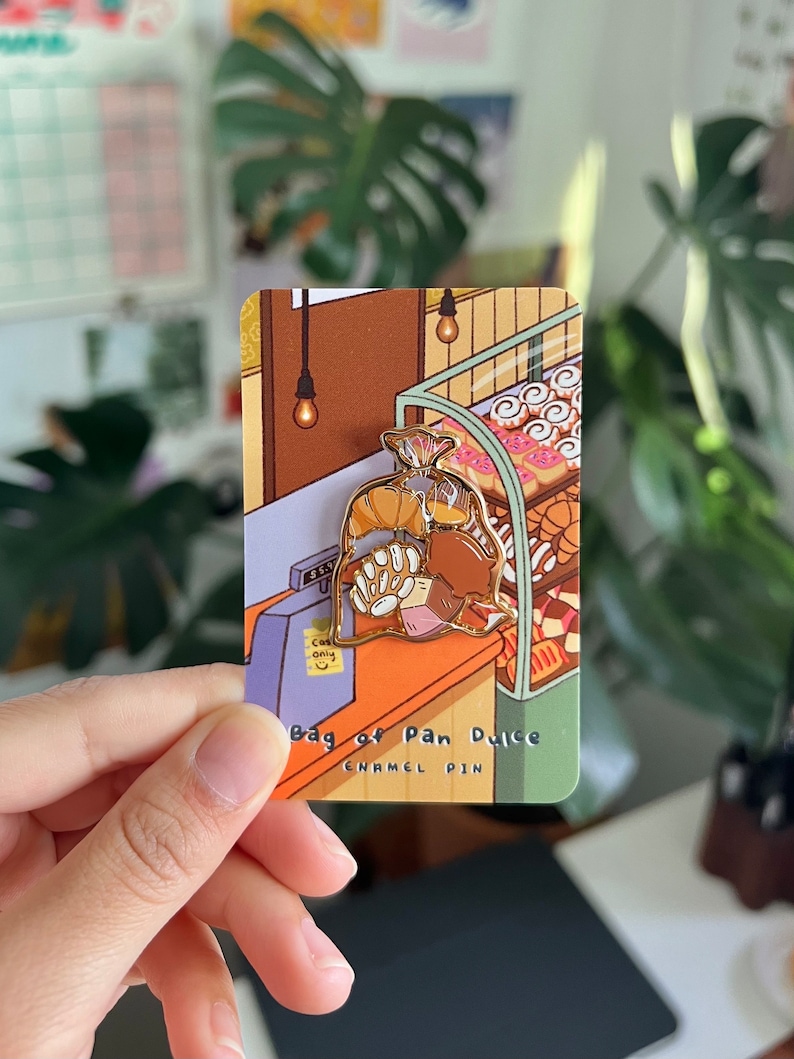 K&ouml;nnte beinhalten: Eine goldene Emaille-Anstecknadel, die einen braunen Papierbeutel zeigt, der mit Pan Dulce gef&uuml;llt ist, einer Art s&uuml;&szlig;em Brot. Die Anstecknadel ist vor einem Comic-Hintergrund einer B&auml;ckerei mit einer Kasse und Regalen mit Geb&auml;ck platziert. Der Text "Bag of Pan Dulce Enamel Pin" ist unter der Anstecknadel gedruckt.
