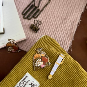 K&ouml;nnte beinhalten: Zwei goldene Emaille-Pins auf einem senfgelben Cordstoff. Ein Pin ist ein wei&szlig;er Stift mit einem blauen Tintenfass und der andere ist eine T&uuml;te mit Geb&auml;ck. Der Text auf dem Stoff lautet "Oh, lolly day!"