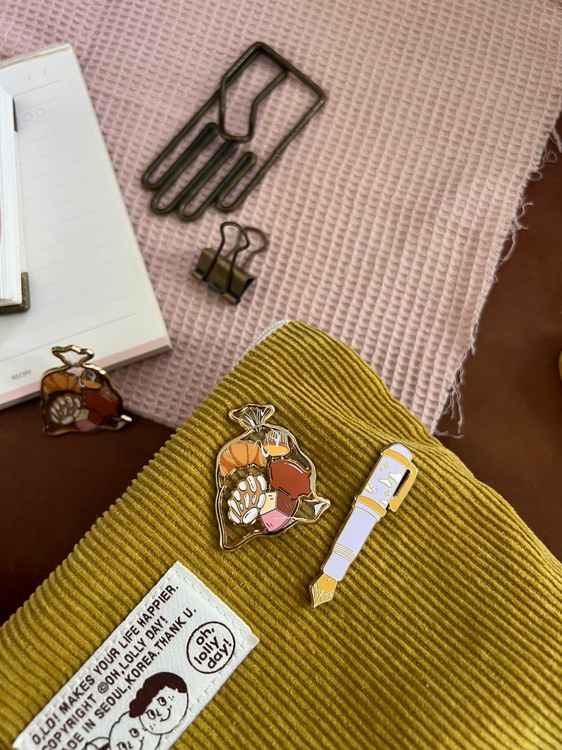 K&ouml;nnte beinhalten: Zwei goldene Emaille-Pins auf einem senfgelben Cordstoff. Ein Pin ist ein wei&szlig;er Stift mit einem blauen Tintenfass und der andere ist eine T&uuml;te mit Geb&auml;ck. Der Text auf dem Stoff lautet "Oh, lolly day!"