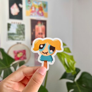 Puede incluir: Una pegatina de dibujos animados de una ni&ntilde;a con cabello rubio y un vestido azul, llorando y derriti&eacute;ndose como un cono de helado. La pegatina est&aacute; sobre un fondo blanco.