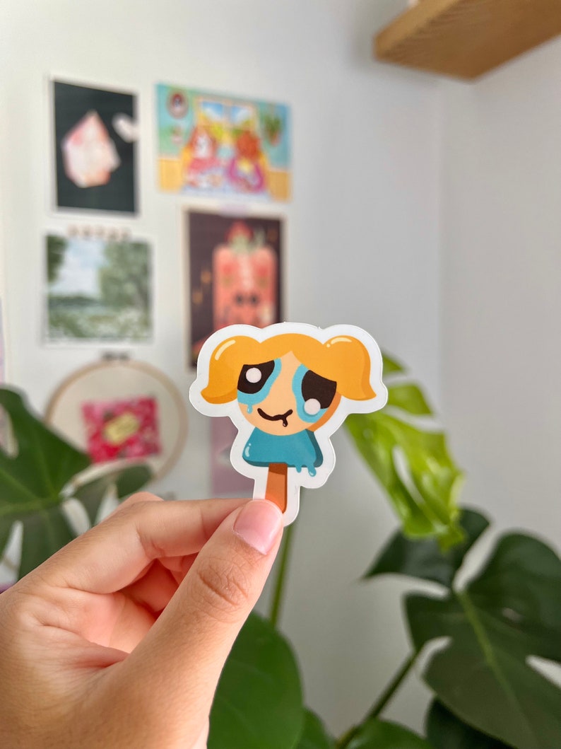 Puede incluir: Una pegatina de dibujos animados de una ni&ntilde;a con cabello rubio y un vestido azul, llorando y derriti&eacute;ndose como un cono de helado. La pegatina est&aacute; sobre un fondo blanco.