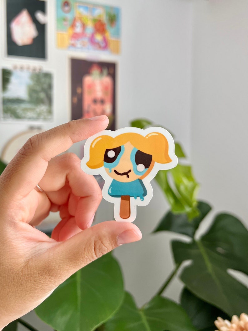 Puede incluir: Una pegatina de una ni&ntilde;a de dibujos animados con cabello rubio y ojos azules. Lleva un vestido azul y tiene una expresi&oacute;n triste en su rostro. Est&aacute; sosteniendo una paleta de hielo en su mano.