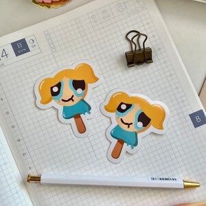 Puede incluir: Dos pegatinas de dibujos animados de una ni&ntilde;a con cabello rubio y ojos azules, vestida con un vestido azul y sosteniendo una paleta de hielo. La ni&ntilde;a est&aacute; llorando y tiene una l&aacute;grima rodando por su mejilla. Las pegatinas est&aacute;n en un cuaderno blanco cuadriculado.