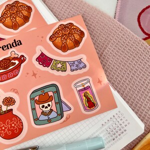 Op de afbeelding: Een set Day of the Dead-thema stickers met een suiker schedel, een pan de muerto, een kaars, een bloempot en een banner. De stickers zijn op een roze achtergrond.